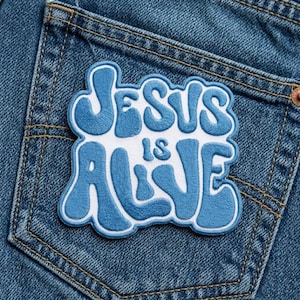 May include: A blue and white embroidered patch with the words "JESUS IS ALIVE" in a bubble font. The patch is sewn onto the back pocket of a pair of blue denim jeans.