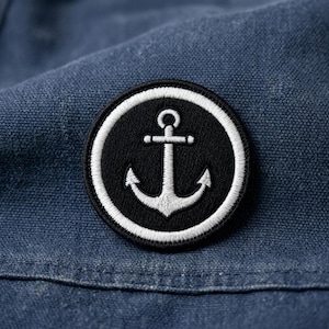 May include: A round, black and white embroidered patch featuring a nautical anchor design. The anchor is white and centered on a black background, surrounded by a white border. The patch is set against a blue fabric.