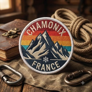 May include: A circular embroidered patch featuring a mountain range in dark blue and white, set against a sunset background of red, orange, and teal stripes. The text reads CHAMONIX at the top and FRANCE at the bottom, with a small white snowflake icon.