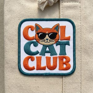 May include: Embroidered patch featuring a cool cat wearing sunglasses. The patch reads "COOL CAT CLUB" in orange and teal lettering, outlined in a teal border. The patch is square with rounded corners and is set against a beige fabric background.