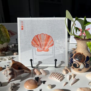 May include: A white canvas with a hand-stitched orange seashell design, mounted on a black hook rack. Various seashells and a decorative vase with greenery are arranged around the canvas, creating a coastal theme.