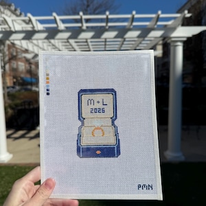 May include: Needlepoint canvas featuring a blue ring box with a gold ring inside. The box has the initials "M-L" and the year "2026" embroidered on it. The canvas is held in front of a white pergola and blue sky.