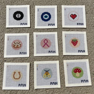 May include: Nine square needlepoint canvases featuring individual designs including an eight ball, evil eye, red heart, cinnamon roll, pink ribbon, strawberry, horseshoe, moth, and mushroom, each with the letters PMN printed in the bottom corner.