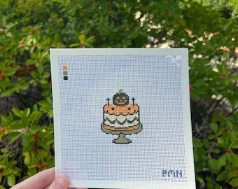 Op de afbeelding: Een vierkant borduurcanvas met een kruissteekontwerp van een oranje taart met een pompoen bovenop een groene taartstandaard. Het canvas bevat een klein kleurenpalet in de linkerbovenhoek en de letters PMN in de rechterbenedenhoek.