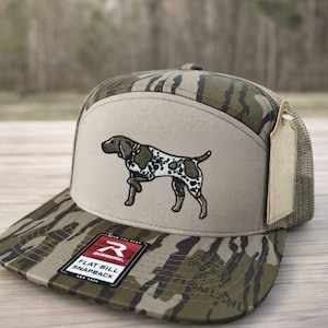 May include: A camouflage flat bill snapback hat with a tan front panel featuring an embroidered dog. The hat has a mesh back and a tag attached. The front of the hat has the text "FLAT BILL SNAPBACK".