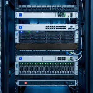 Può includere: Un rack server pieno di hardware di rete. Il rack contiene più server, switch e unità di distribuzione dell'alimentazione. I dispositivi sono neri e argentati, con varie porte e spie luminose. I cavi sono ben organizzati.