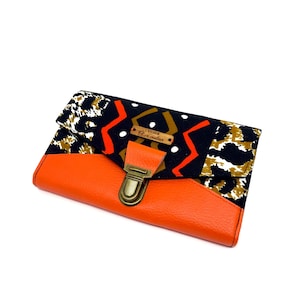 May include: An orange leather clutch with a patterned flap. The flap features a black background with white, orange, and gold geometric shapes. A small leather tag is attached to the flap. The clutch has a metal clasp.