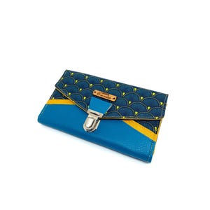 May include: A blue wallet with a geometric pattern on the flap. The flap features a navy blue fan-shaped design with yellow accents. The wallet has a silver clasp and a small wooden tag.
