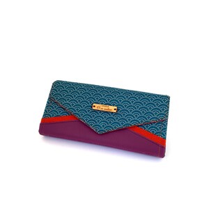 May include: A rectangular wallet with a teal wave pattern flap and a deep purple body. A thin red stripe accents the flap's edge. A small wooden tag with text is affixed to the flap. The wallet is approximately 7 inches long.