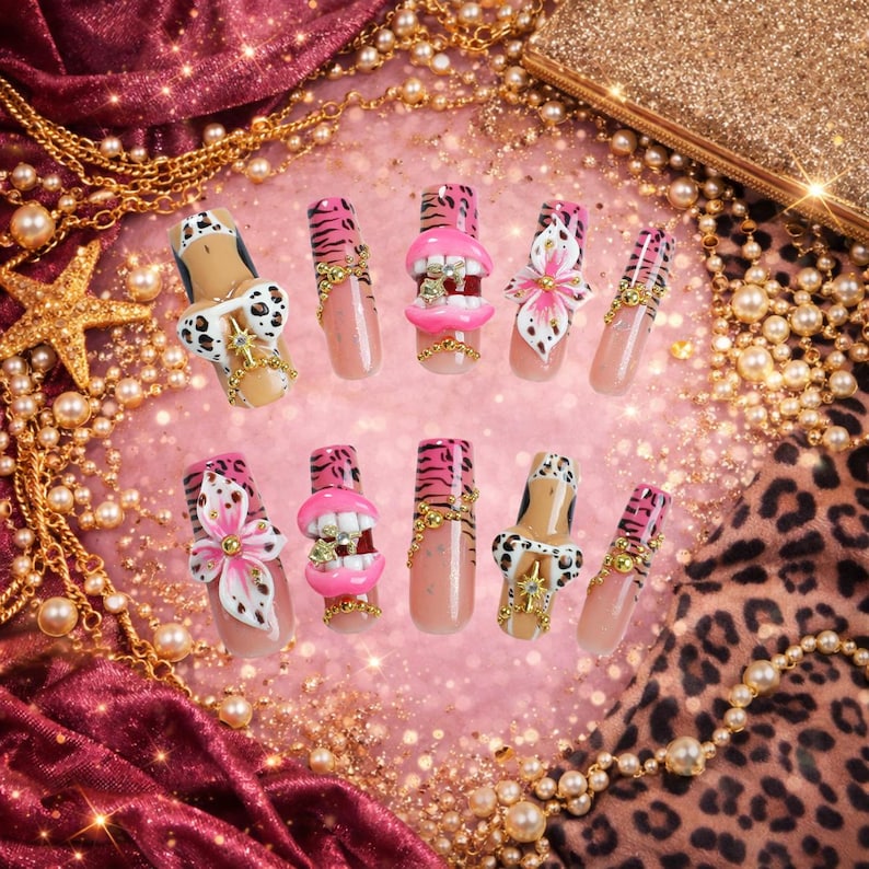 May include: A set of ten press-on nails featuring a variety of designs including pink tiger stripes, 3D pink lips with white teeth, white and black leopard print patterns, pink floral accents, and gold-toned chain embellishments with small crystals.