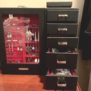 May include: A black jewelry box with a red velvet interior and a glass door displaying various earrings, rings, and necklaces. The box has multiple drawers for additional storage. The jewelry box is on a wooden surface.