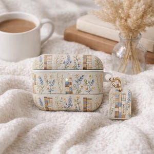 May include: A beige AirPod case and matching keychain with embroidered floral and book designs. The case and keychain feature blue, yellow, and brown accents. A white mug of coffee and a vase of dried flowers are in the background.