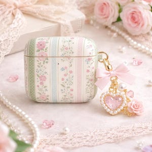 May include: A floral-patterned AirPod case with a pink bow and heart-shaped charm. The case features vertical stripes in green, pink, and blue, adorned with pink flowers. A gold-colored clasp holds the charm, which includes a pearl and a small pink rose.
