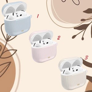 May include: Three open AirPod cases in pastel colors: light blue, pink, and cream. Each case has vertical stripes and contains white AirPods. The cases are labeled with "SM" and are set against a neutral background with floral accents.
