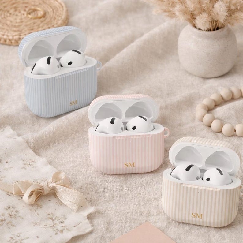 May include: Three open AirPod cases in pastel colors: light blue, pink, and beige. Each case has vertical stripes and the initials "SM" on the front. The AirPods inside are white with black accents. The cases are on a neutral-toned fabric.