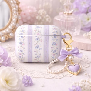 May include: A white and lavender floral patterned case for wireless earbuds. The case has a gold-colored clasp and is adorned with a lavender bow, pearl beads, and a heart-shaped charm. The overall aesthetic is delicate and feminine.