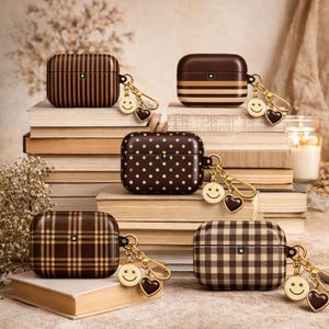 May include: Five brown patterned AirPod Pro cases with gold-colored keychains. The cases feature various designs, including stripes, polka dots, and plaid. Each keychain has a smiley face charm and a heart charm. The cases are displayed on a stack of books.