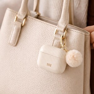 May include: A cream-colored, striped AirPods case with the initials "SM" is attached to a matching handbag. A small, fluffy pom-pom charm hangs from the case. The handbag has gold-colored hardware.