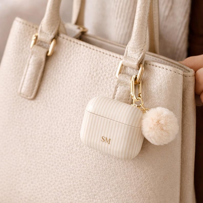 May include: A cream-colored, striped AirPods case with the initials "SM" is attached to a matching handbag. A small, fluffy pom-pom charm hangs from the case. The handbag has gold-colored hardware.