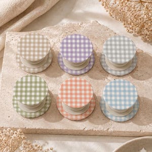 May include: Six colorful phone grips with a gingham pattern in shades of beige, lavender, gray, green, coral, and blue. The grips are round with a white base and are arranged on a stone surface.