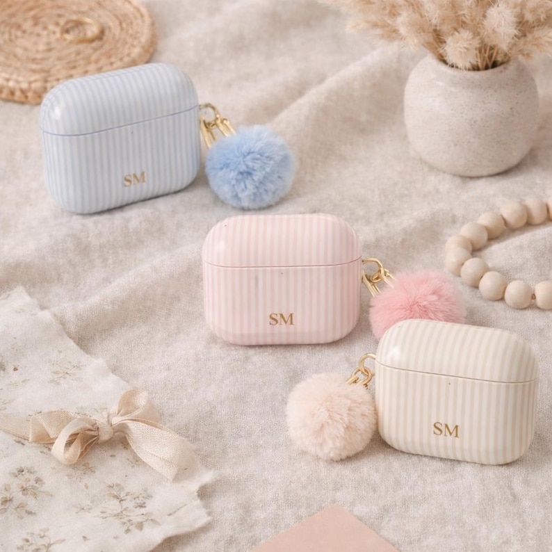 May include: Three striped AirPod cases in pastel colors: light blue, pink, and cream. Each case has a gold-colored keychain with a fluffy pom-pom and the initials "SM". The cases are on a soft, neutral-colored surface.