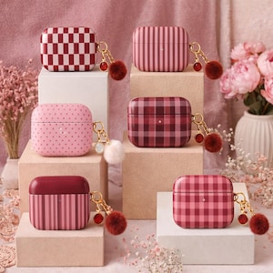 May include: A collection of patterned AirPod cases in various designs, including checkered, striped, and polka dot patterns. Each case is in shades of pink and burgundy, and features a gold-colored keychain with a decorative charm.