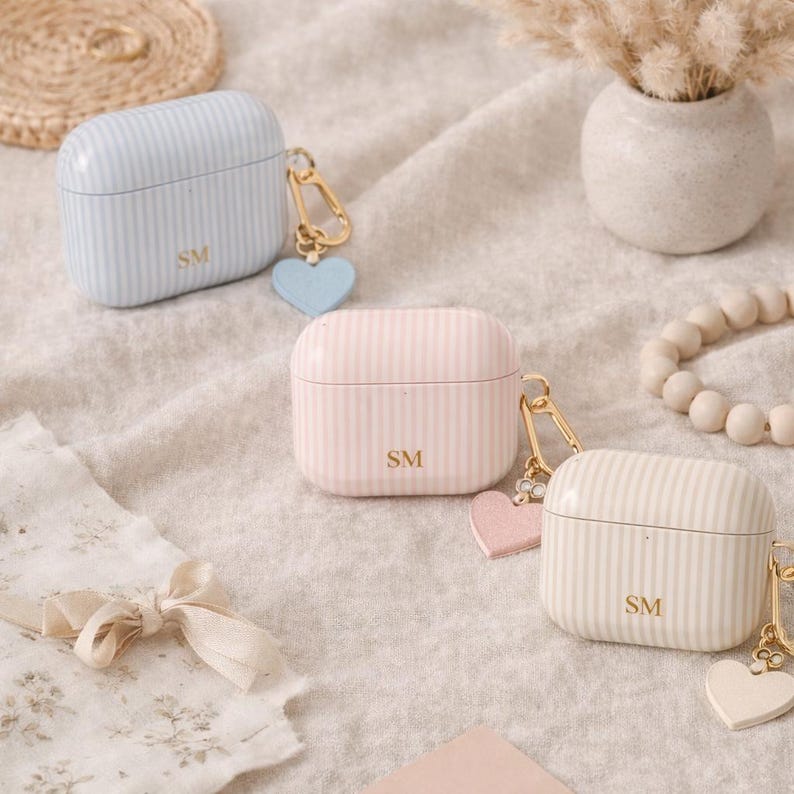 May include: Three striped AirPod cases in pastel colors: light blue, pink, and off-white. Each case has a gold-colored clasp, a heart-shaped charm, and the initials "SM". The cases are on a neutral-toned fabric.