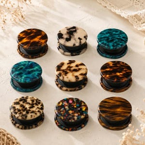 May include: Nine round phone grips in various colors and patterns. The grips have a tortoiseshell design in shades of brown, beige, and black, as well as teal and multicolored patterns. Each grip has a black base and a collapsible design.