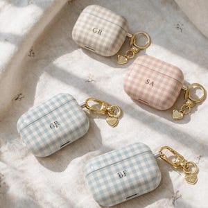 May include: Four personalized AirPod cases in pastel colors: beige, pink, and two shades of blue. Each case has a gingham pattern and gold-colored hardware with a heart charm. The cases are marked with initials.