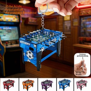 May include: A blue foosball table keychain with the name "ANTHONY" on the front. The keychain is made of 2D flat acrylic and has a thickness of 0.15 inches. The design includes soccer balls and the text "2D flat Acrylic. Please peel off the protective film behind before use."