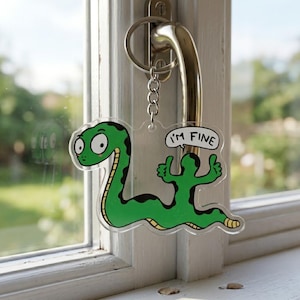 May include: A clear acrylic keychain featuring a cartoon green snake with black spots and a yellow underbelly. The snake has wide eyes and is giving two thumbs up. A speech bubble above the snake contains the black text I'M FINE.