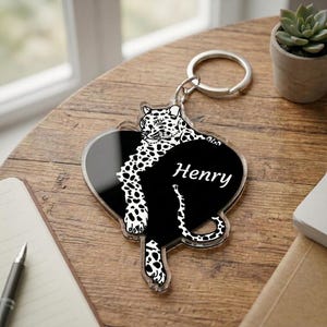 May include: A clear acrylic keychain featuring a black heart-shaped design with a white leopard illustration and the name Henry printed in white script. The keychain includes a silver-toned metal split ring for attachment to keys or bags.