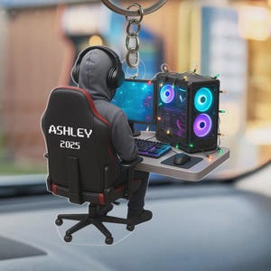 May include: A gaming-themed keychain featuring a gamer seated at a desk with a computer setup. The chair has "ASHLEY 2025" printed on it. The computer case is decorated with colorful lights, and the gamer wears headphones.