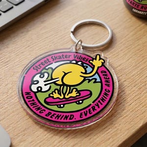 May include: A clear acrylic keychain with a cartoon character on a skateboard. The design features a yellow character with a raised middle finger, riding a pink skateboard on a green background. Text includes "Street Skater Vibes" and "Nothing Behind. Everything Ahead."