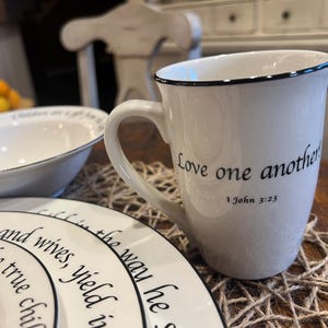 May include: A white ceramic mug with a black rim and handle, featuring the black printed text Love one another! 1 John 3:23. The mug is positioned next to white plates and a bowl, all decorated with black text and thin black rim detailing.