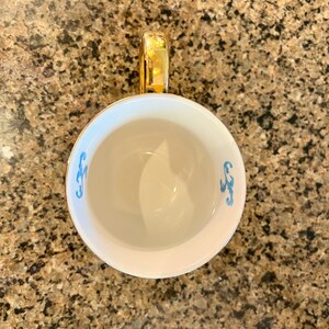 May include: White teacup with a gold handle and rim. The interior is white, and the exterior features a light blue decorative design. The teacup is set against a speckled brown background.