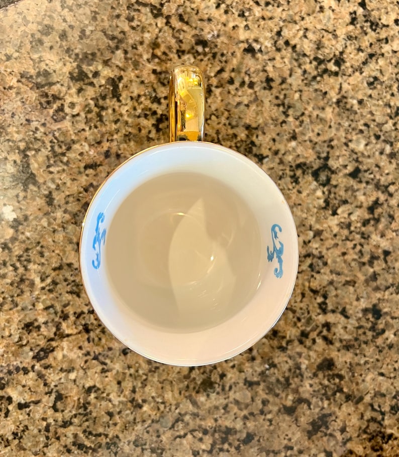 May include: White teacup with a gold handle and rim. The interior is white, and the exterior features a light blue decorative design. The teacup is set against a speckled brown background.