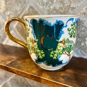 May include: A ceramic mug with a gold handle and rim. The mug features a dark teal background with a gold letter "C" and illustrations of chameleons and foliage. The design is framed by a white and blue border.
