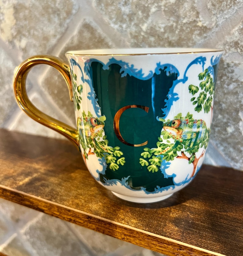 May include: A ceramic mug with a gold handle and rim. The mug features a dark teal background with a gold letter "C" and illustrations of chameleons and foliage. The design is framed by a white and blue border.
