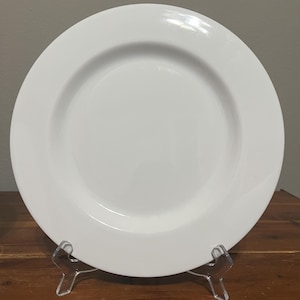 May include: A white ceramic plate with a wide rim, displayed on a clear plastic stand. The plate has a smooth, glossy surface and a simple, elegant design. The plate is round and appears to be approximately 10 inches in diameter.