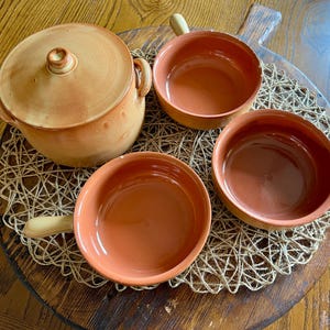 May include: A set of terracotta cookware, including a lidded pot and three handled bowls. The items are arranged on a wooden cutting board with a woven placemat. The cookware has a rustic, earthy aesthetic.