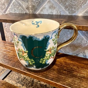 May include: A decorative ceramic mug with a gold handle and rim. The mug features a green panel with the letter "C" and a floral and bird design. The mug is set on a wooden shelf.