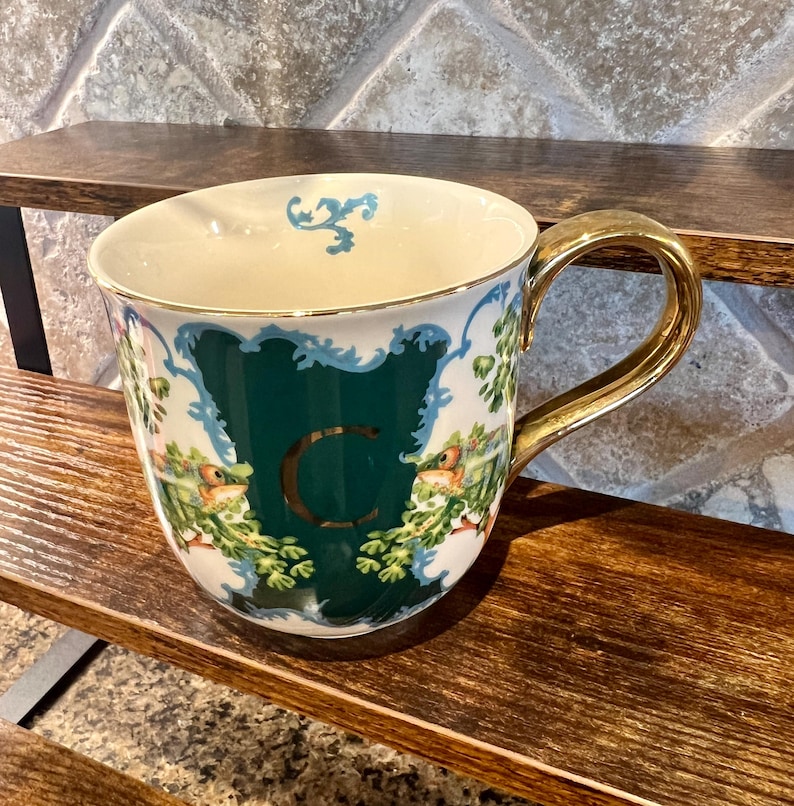 May include: A decorative ceramic mug with a gold handle and rim. The mug features a green panel with the letter "C" and a floral and bird design. The mug is set on a wooden shelf.