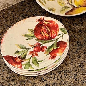 May include: A stack of white ceramic plates with a pomegranate design. The plates feature a watercolor illustration of a ripe pomegranate with red fruit, green leaves, and small red berries. The plates are stacked on a brown surface.