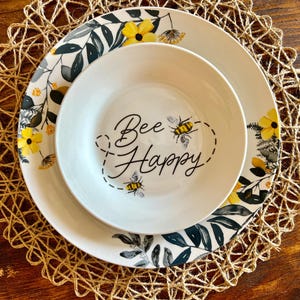 May include: A white ceramic bowl and plate set featuring a black and yellow bee illustration with the text Bee Happy. The plate is decorated with a botanical pattern of dark green leaves and yellow flowers, while the bowl features two bees.