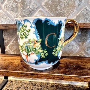 May include: A white ceramic mug with a gold handle and rim. The mug features a dark teal background with a white and blue ornate border. A large gold letter "C" is centered on the mug, with a chameleon and foliage design.