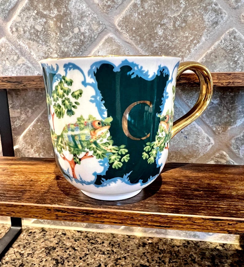 May include: A white ceramic mug with a gold handle and rim. The mug features a dark teal background with a white and blue ornate border. A large gold letter "C" is centered on the mug, with a chameleon and foliage design.