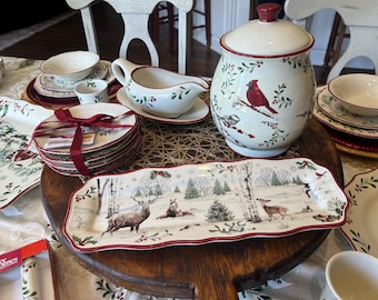 May include: A collection of cream-coloured ceramic dinnerware featuring holiday motifs including red cardinals, holly, and winter forest scenes with deer. The set includes a lidded canister, a gravy boat, a rectangular serving platter, and stacked plates.