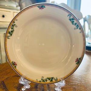 May include: Cream-colored ceramic plate with a decorative border. The border features a yellow band with a black outline, adorned with green vines and red floral accents. The plate is displayed on a clear plastic stand.