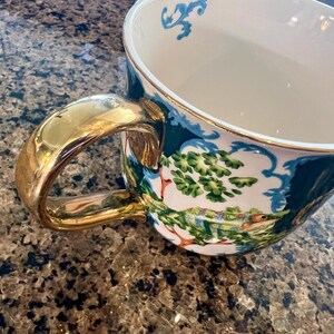 May include: A white ceramic teacup with a gold handle and rim. The cup features a detailed landscape scene with green trees and a colorful chameleon. Blue decorative accents are on the rim.